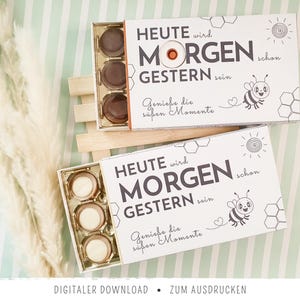 Könnte beinhalten: Zwei rechteckige weiße Schachteln mit Pralinen, bedruckt mit dem Text HEUTE wird MORGEN schon GESTERN sein und Genieße die süßen Momente, ergänzt durch eine Bienenillustration, eine Sonne und Wabenmuster auf der Verpackung.