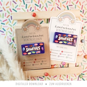 May include: Two rectangular cards featuring a rainbow and flower illustration at the top with the text "AUF EIN kunterbuntes RENTNERLEBEN" and "MIT GANZ VIEL". Each card holds a small box of Smarties mini candies, with printed lists of German words below.