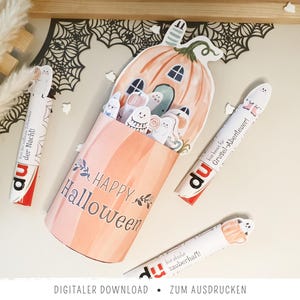 Könnte beinhalten: Ein zylindrischer Papierbehälter mit Kürbis-Illustration und dem Text Happy Halloween, gefüllt mit kleinen Geister-Ausschnitten. Drei weitere Papierhüllen mit Geister-Illustrationen und dem Text du sind um den Behälter herum angeordnet.