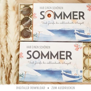 May include: Two rectangular gift boxes containing round chocolates, featuring a watercolour illustration of a boat and beach umbrella. The text reads HAB EINEN SCHÖNEN SOMMER Und genieße die wohlverdiente Auszeit. One box holds dark chocolates, the other white.