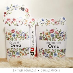 May include: Two white paper gift boxes with floral designs and the words "FÜR DIE BESTE Oma" and "MIT LIEBE FÜR Oma". The boxes are decorated with watercolor flowers in pink, blue, and yellow. A floral wreath is on top.