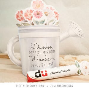 May include: A watercolor-style watering can with a handle and spout, filled with paper flowers in shades of pink and white. The can has text that reads "Danke, dass du mir beim Wachsen geholfen hast." A candy bar with the text "du schenkst Freude" is also present.