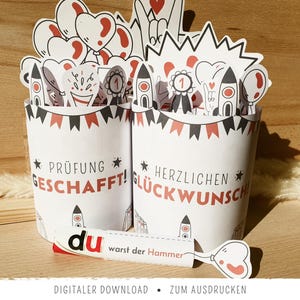 May include: Two white paper cups with red and black accents, decorated with rockets, balloons, and banners. One cup says "Prüfung Geschafft!" and the other says "Herzlichen Glückwunsch". A small card reads "du warst der Hammer".