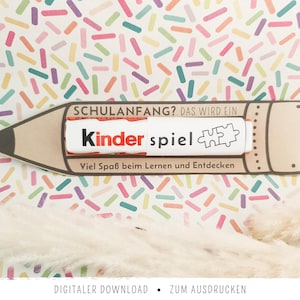 Op de afbeelding: Een potloodvormige papieren kaart met de tekst SCHULANFANG? DAS WIRD EIN Kinder spiel en Viel Spaß beim Lernen und Entdecken, met een Kinder-chocoladereep bevestigd in het midden van het potloodontwerp.