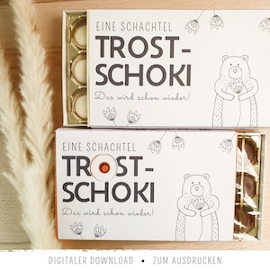Può includere: Due scatole rettangolari bianche etichettate EINE SCHACHTEL TROST-SCHOKI Das wird schon wieder! con un disegno a linee nere di un orso che tiene un fiore. Le scatole sono aperte, rivelando vassoi di plastica dorata contenenti cioccolatini rotondi.