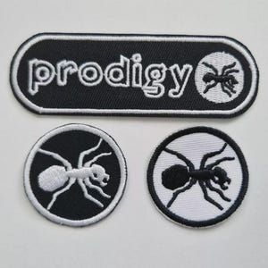 Pode incluir: Três patches bordados. O patch superior é um retângulo preto com cantos arredondados, a palavra "prodigy" em branco e um gráfico de formiga branca. Dois patches circulares apresentam gráficos de formigas, um branco sobre preto, o outro preto sobre branco.