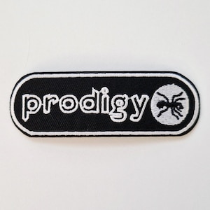 Può includere: Una toppa ricamata rettangolare con angoli arrotondati, sfondo nero e bordo bianco. La parola prodigy è ricamata in lettere minuscole bianche accanto a un emblema circolare bianco contenente un'illustrazione grafica di una formica nera.