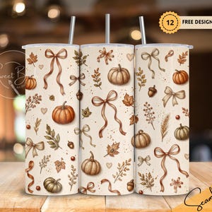 May include: Three cylindrical tumblers with straws featuring a cream-coloured background decorated with a repeating pattern of orange and brown pumpkins, tan ribbons, autumn leaves, acorns, and wheat stalks. The text 12 FREE DESIGNS is visible.