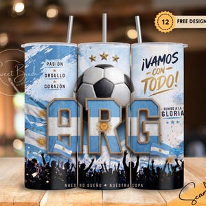 May include: A cylindrical tumbler featuring a soccer theme with the Argentine flag colors, a soccer ball, and the text "ARG", "&iexcl;VAMOS CON TODO!", "PASI&Oacute;N ORGULLO CORAZ&Oacute;N", "RUMBO A LA GLORIA", and "NUES SUE&Ntilde;O NUESTRA COPA" with a straw and a lid.