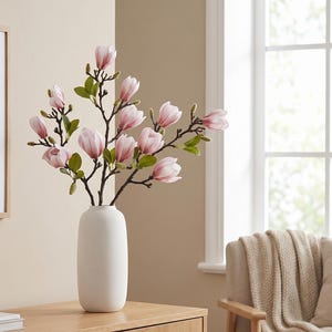 Op de afbeelding: Een hoge, cilindrische matwitte vaas bevat een tak met kunstmatige magnolia bloemen. De bloemen zijn zachtroze met witte centra, vergezeld van donkerbruine stelen en kleine groene bladeren. Het arrangement staat rechtop en in balans.