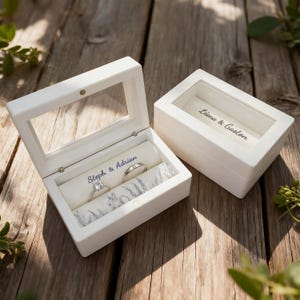 May include: Two white ring boxes with clear lids on a wooden surface. One is open, showing a ring and the text "Steph & Adrian" on a fabric roll. The closed box has the text "Liana & Gaston" visible.