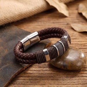 Op de afbeelding: Een armband van donkerbruin gevlochten leer met een gepolijste magnetische sluiting van zilverkleurig metaal en drie decoratieve zilverkleurige metalen accenten, waaronder een centrale kraal met stippelpatroon en twee gladde ringen.
