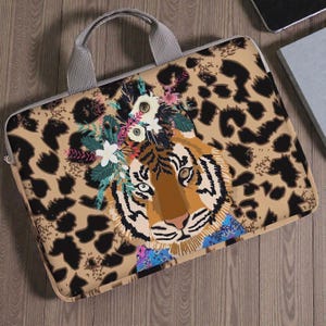 Può includere: Una custodia per laptop con un'illustrazione di tigre con una corona floreale, su uno sfondo con stampa leopardata beige. La custodia ha un manico grigio e una chiusura a zip. La tigre ha strisce arancioni e nere, con un colletto blu e rosa.