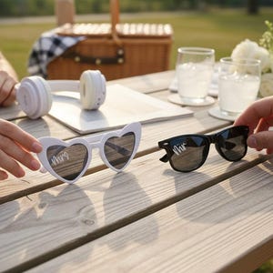 Pode incluir: Cena de piquenique com óculos de sol brancos em forma de coração com "Mrs" e óculos de sol pretos com "Mr". Um par de fones de ouvido brancos e uma cesta de piquenique estão na mesa de madeira.