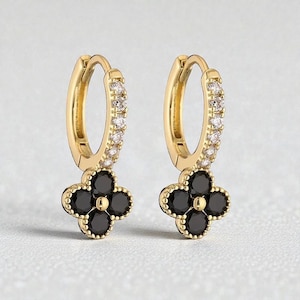 Peut inclure: Une paire de boucles d'oreilles créoles dorées. Chaque boucle d'oreille est ornée d'un charm en forme de trèfle noir à quatre feuilles et de petits pierres précieuses transparentes le long de l'anneau. Les boucles d'oreilles sont présentées sur un fond clair.