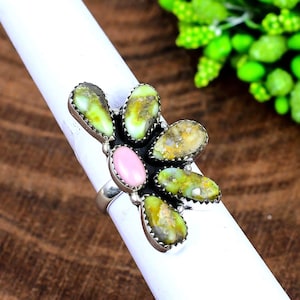 May include: A silver ring featuring a cluster of gemstones. The ring has a central pink oval stone surrounded by six teardrop-shaped green and yellow stones. The gemstones are set in a silver metal setting.