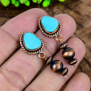 May include: A pair of turquoise and copper earrings. Each earring features a teardrop-shaped turquoise stone set in a twisted copper frame. Below the turquoise stone hang three copper beads. The earrings are designed for pierced ears.