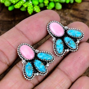 May include: A pair of stud earrings featuring a central oval pink stone surrounded by three smaller oval turquoise stones. The stones are set in a silver metal frame with a beaded edge detail, creating a cluster design for each earring.