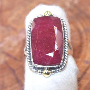 Puede incluir: Anillo rectangular de rubí con banda de plata y detalles dorados. El rubí es de color rojo intenso y tiene un corte facetado. El anillo tiene un diseño de cuerda trenzada alrededor de la gema.