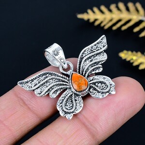 May include: A silver-colored pendant in the shape of a butterfly with an orange gemstone center. The pendant has detailed wing and body designs. The bail features a sunburst design. The pendant is held by a person's fingers.
