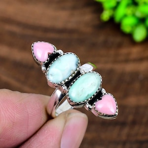 May include: A silver ring with three oval, pale blue gemstones and two pink heart-shaped stones. The ring has an intricate design and is held by a hand against a blurred background.