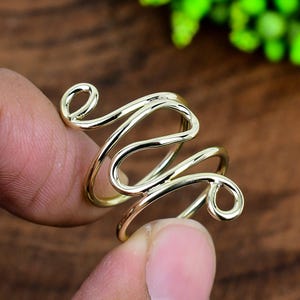 Puede incluir: Un anillo dorado con un diseño único y fluido. El anillo presenta una serie de líneas curvas interconectadas que se envuelven alrededor del dedo. El diseño del anillo incluye dos espirales circulares. El anillo se sostiene entre dos dedos.