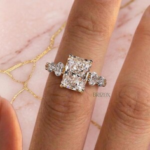 Könnte beinhalten: Ein goldener Ring mit einem großen, rechteckig geschliffenen Diamanten und kleineren Seitensteinen. Der Ring wird auf einem Finger vor einem rosa-weißen Marmorhintergrund gezeigt. Der Markenname "BRIZON" ist auf dem Ring sichtbar.