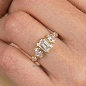 May include: A gold ring featuring an emerald-cut diamond center stone, flanked by smaller round diamonds. The ring is set in a yellow gold band and is worn on a finger.