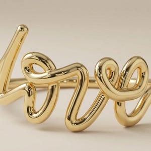 Może przedstawiać: Złoty pierścionek w kształcie słowa "love". Pierścionek wykonany jest z błyszczącego, gładkiego metalu i ma kursywę. Słowo "love" jest głównym elementem projektu, a litery połączone są w ciągłą linię.