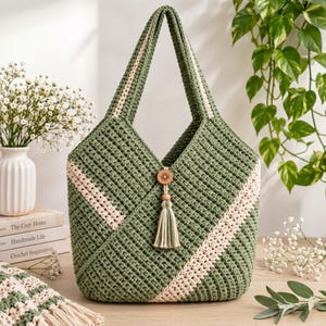 May include: A sage green crochet tote bag featuring diagonal cream-colored stripes, two integrated handles, and a central wooden button closure with a matching sage green and cream-colored yarn tassel hanging from the front center.