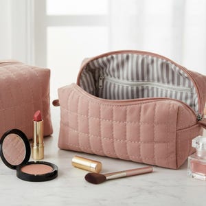 May include: A pink quilted cosmetic bag with a zipper, open to show a striped interior. Another bag is visible, alongside makeup items such as lipstick, blush, a brush, and a compact mirror. The items are arranged on a white surface.