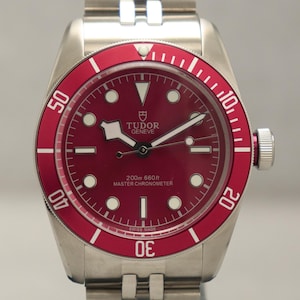 Puede incluir: Reloj de pulsera de acero inoxidable con bisel y esfera rojos. El reloj tiene una pulsera plateada y el texto "TUDOR GENEVE" en la esfera. Es un Master Chronometer y resistente al agua hasta 200m. La pulsera es plateada.