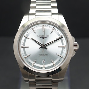 May include: A silver-toned Longines Conquest Automatic watch with a metallic silver dial, silver hour markers, and silver hands. The watch features a date window at the six o'clock position and a stainless steel link bracelet with a folding clasp.