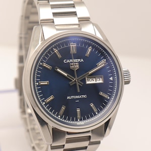Può includere: Un orologio da polso in acciaio inossidabile color argento con quadrante blu profondo, indici e lancette argentati. Il quadrante riporta la scritta Carrera, Tag Heuer, Automatic e Swiss, con una finestrella per giorno e data Mon 13.