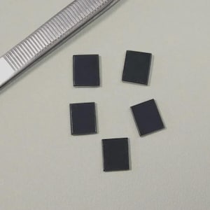 Puede incluir: Cinco pequeños componentes cuadrados de color gris oscuro están dispersos sobre una superficie gris claro. Un par de pinzas plateadas están colocadas cerca de los componentes. Los componentes parecen ser piezas electrónicas.