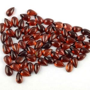 Può includere: Una collezione di gemme lucide a forma di goccia, nei toni del rosso e del marrone. Le gemme sono sparse su una superficie bianca, mostrando la loro qualità traslucida e le loro varie sfumature. Adatto per la creazione di gioielli.