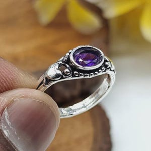 Könnte beinhalten: Ein Silberring mit einem zentralen, oval geschliffenen violetten Edelstein in einer dekorativen Metallfassung. Das Band weist aufwendige perlenartige Verzierungen und einen kleinen herzförmigen Akzent an der Seite auf.