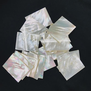 May include: A collection of iridescent, square mother-of-pearl shell pieces. The shells have a pearlescent sheen with hints of pink, green, and blue. The pieces are arranged on a dark background.