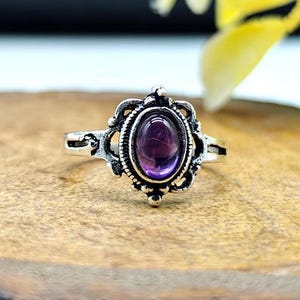 Pode incluir: Um anel de tom prateado com uma pedra preciosa roxa de formato oval no centro. A banda de metal apresenta um padrão de filigrana decorativo e intrincado ao redor da pedra, com um acabamento polido no engaste e um aspeto oxidado mais escuro.