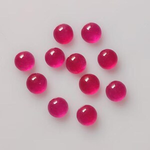 May include: A collection of eleven round, polished, deep pink gemstones. Each gem has a smooth, reflective surface, with a bright highlight. The gems are scattered on a light gray surface, creating a visually appealing arrangement.