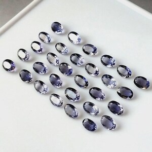 Peut inclure: Une collection de pierres précieuses ovales, bleu-violet, disposées sur une surface blanche. Les pierres polies reflètent la lumière, mettant en valeur leur clarté et leur couleur. L'arrangement suggère une présentation de pierres précieuses.