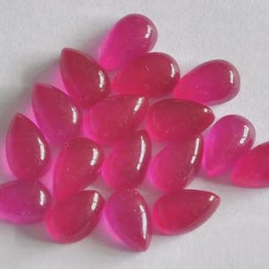 May include: A collection of loose, teardrop-shaped gemstones in a vibrant, translucent fuchsia pink color. Each stone features a smooth, polished cabochon surface with a rounded top and a pointed end, reflecting light across their curved surfaces.