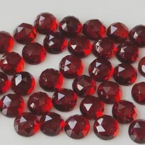 Puede incluir: Una colección de gemas facetadas, redondas, de color rojo oscuro. Las gemas están dispersas sobre una superficie blanca, cada una mostrando múltiples facetas reflectantes. El color es un rojo intenso y rico, con ligeras variaciones de tono.