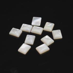 May include: A collection of ten rectangular, off-white mother-of-pearl beads. Each bead has rounded corners and a lustrous, iridescent surface. The beads are scattered against a dark background, showcasing their natural beauty and potential for jewelry making.