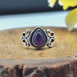 Könnte beinhalten: Ein Silberring mit einem zentralen, birnenförmigen violetten Amethyst-Edelstein, der in eine gedrehte Kordelbordüre eingefasst ist. Die Ringschiene zeigt auf beiden Seiten des Steins komplizierte, durchbrochene Schnörkelmuster in symmetrischem Design.