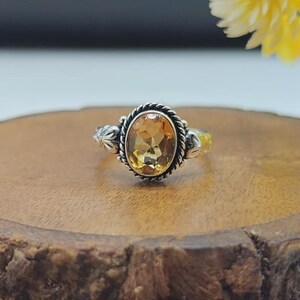 Puede incluir: Un anillo de plata con una piedra preciosa de citrino amarillo de corte ovalado engastada en un borde de metal con diseño de cuerda trenzada. La banda del anillo muestra detalles grabados decorativos a ambos lados, enmarcando la piedra central.