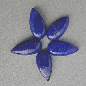 Peut inclure: Cinq pierres précieuses de lapis-lazuli polies en forme de goutte d'eau, disposées en étoile. Les pierres bleu foncé présentent des éclats d'or et d'argent et une finition brillante. Les pierres sont posées sur une surface gris clair.