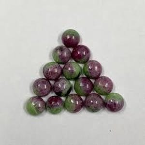 Puede incluir: Una pirámide de piedras preciosas redondas, cada una con una mezcla de verde y rojo intenso. Las piedras tienen un acabado pulido y están dispuestas sobre un fondo liso gris claro. Las piedras son esféricas.