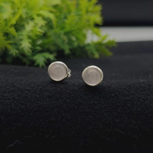 Op de afbeelding: Een paar ronde oorknopjes met gladde, lichtroze rozenkwarts cabochons gezet in gepolijste, zilverkleurige metalen zettingen. De ronde stenen hebben een zachte, doorschijnende uitstraling en zijn gecentreerd in de metalen frames.