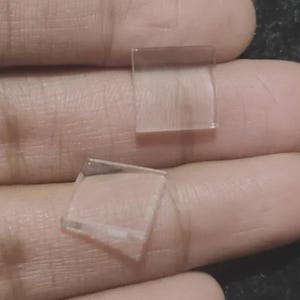 以下が含まれることがあります： 2つの小さく透明な正方形のガラス片。1つは直立し、もう1つは角度がついており、どちらも肌色の表面に置かれています。ガラスは透明に見え、エッジが見えます。